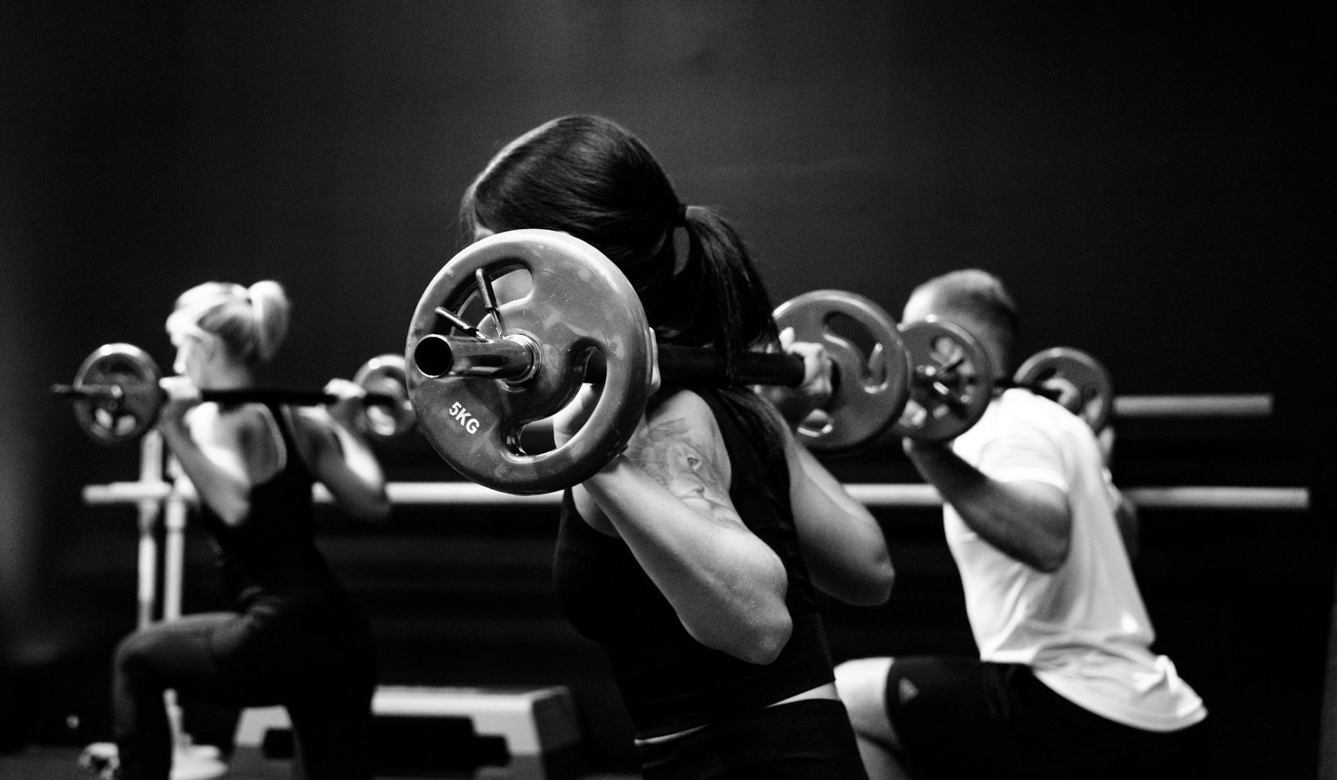Woman in forefront, women and man in background, each with barbell on shoulders facing left in lunge position